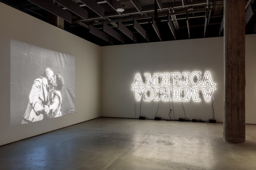 Two gallery walls: the wall on the left features a projection of a vintage black and white film of a Black couple dressed in Victorian clothing kissing in front of a black cloth; on the wall on the right is a neon sign that reads, "America" twice — one facing right side up and the other upside down. 