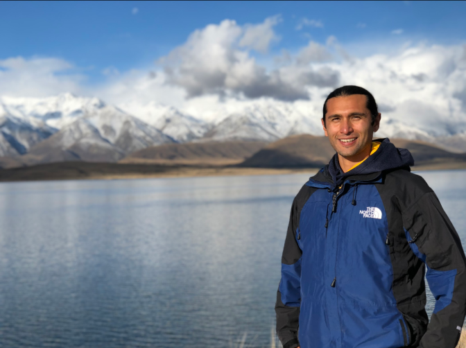 A man in a blue jacket smiles in front of a lake.