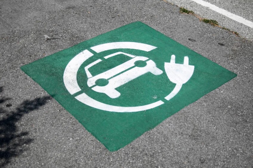 A green and white, square-shaped sign is painted on the pavement with a symbol of a car and an electric cord wrapped around it.