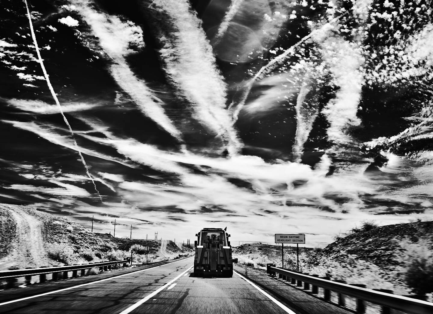 A black and white photo of a truck driving on a desert road against a sky with spraying clouds.