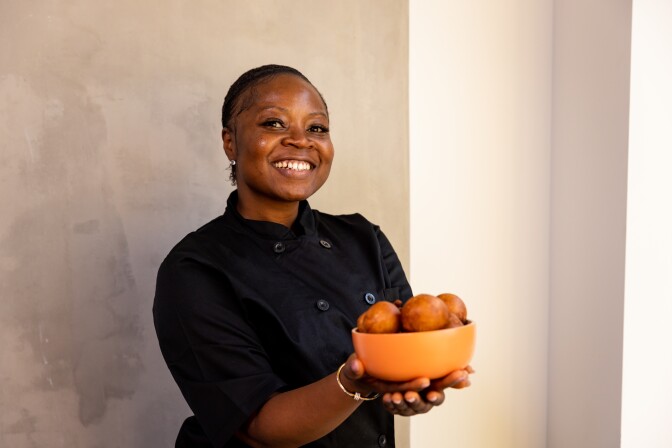 Opportune holds an orange bowl filled with golden brown puffs, each the size of a tennis ball. Opportune is wearing a black chef's shirt and smiles at the camera.