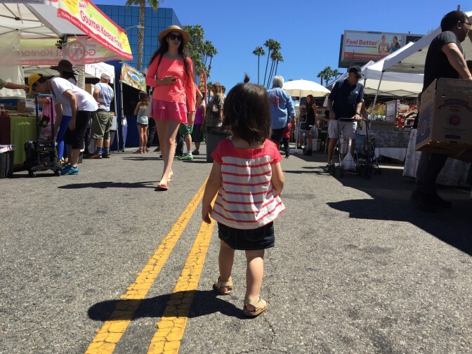 A small child walks down an aisle of farmer's market vendors. The child is wearing a striped pink and white shirt and tan sandals. A clear blue sky stretches above the market and palm trees dot the skyline.