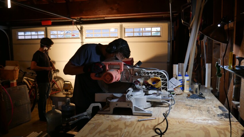 A person hunches over a table saw to cut wood pieces in a garage. Behind them, another person is standing over cut pieces of PVC pipes and wood. 