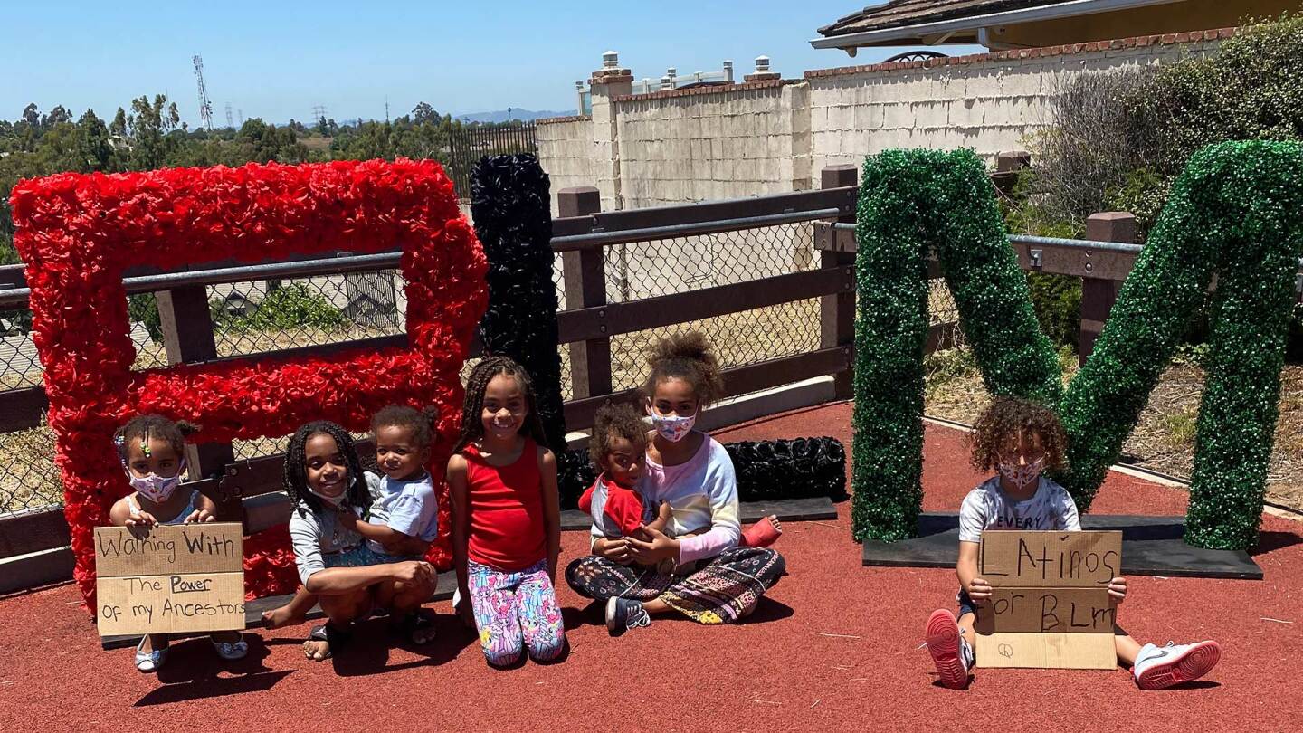 Several children sit in front of a set of large letters spelling out BLM (Black Lives Matter).