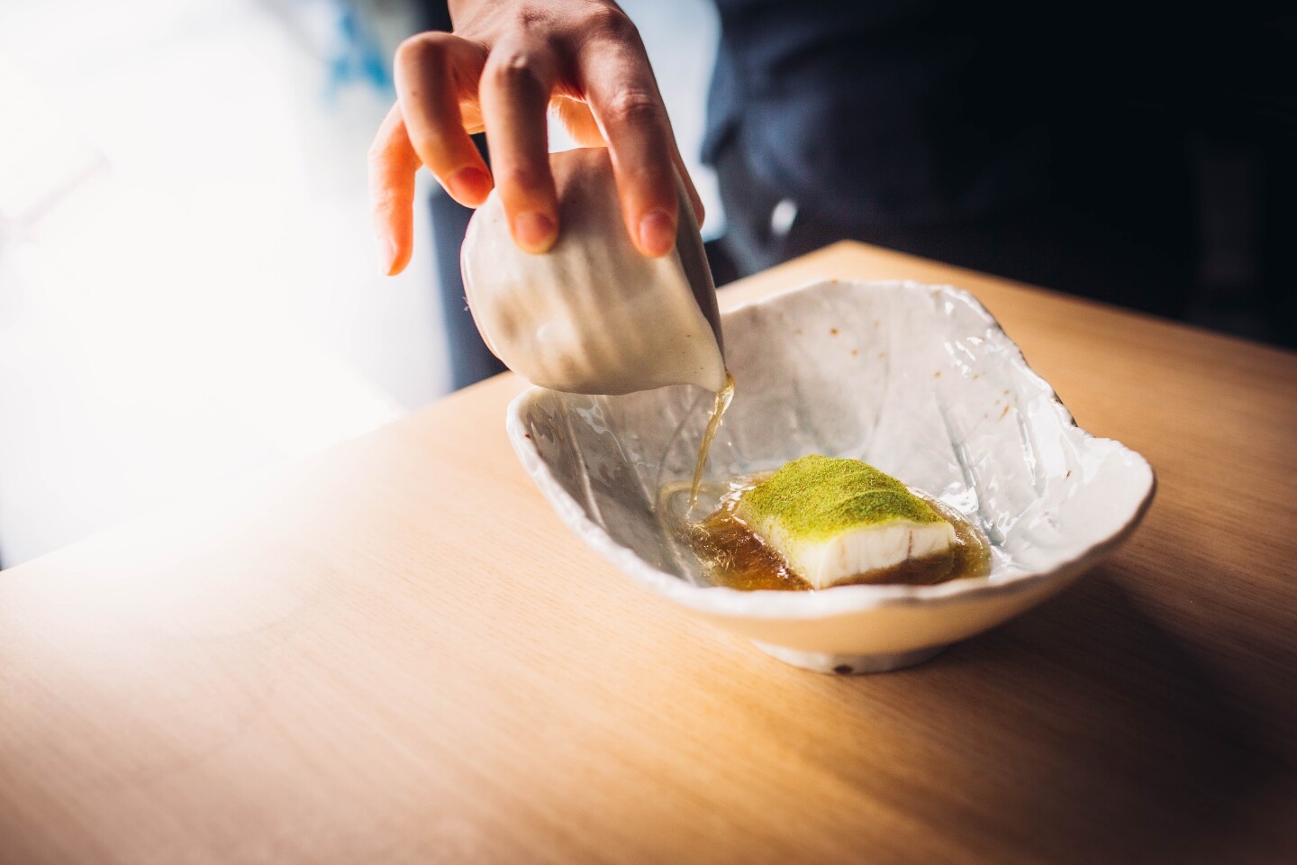 An asymmetrical ceramic dish holds a small, bite-sized piece of white steamed fish sitting in a thin, broth-y sauce. The fish is topped with a fine green powder. Additionally, someone is pouring more of the sauce from a small ceramic container.