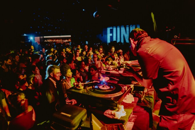 A densely packed crowd listens and watches a performance on a stage that is mostly off-camera. At the forefront is a performer hunched over turntables. The crowd is lit by warm orange and green lights from the stage.