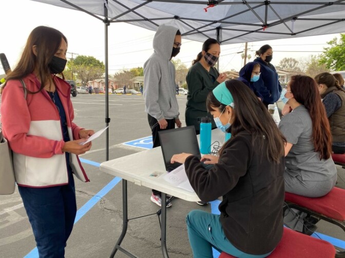 A woman in a halftone pink and white jacket presents stands over a plastic foldable table as she presents her ID to a woman sitting at the table.