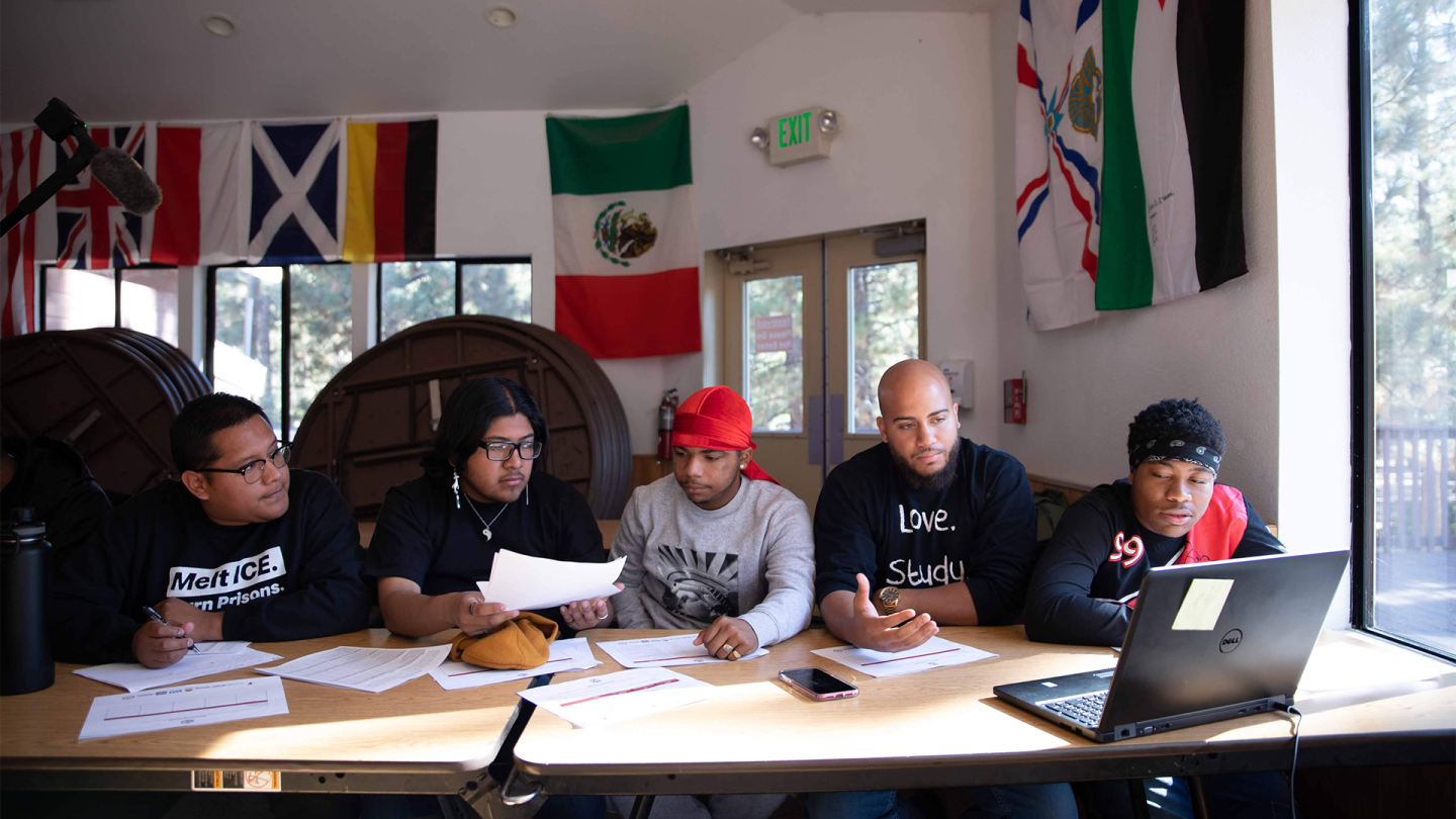 5 men sit at a table in front of a laptop