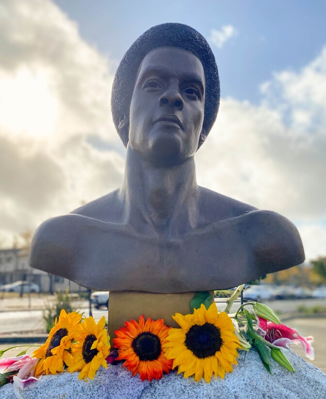 A bronze bust of Huey P. Newton is mounted on a boulder with flowers at the base of the bust. The sun shines behind the clouds beyond.