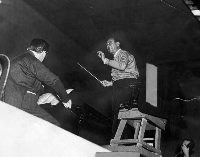A black and white photo taken from below of composer Arnold Schoenberg conducting. He is holding a truncheon and waving as he faces an orchestra, which is out of frame. Sylvain Noack, the concertmaster, is visible on the left, playing the violin.