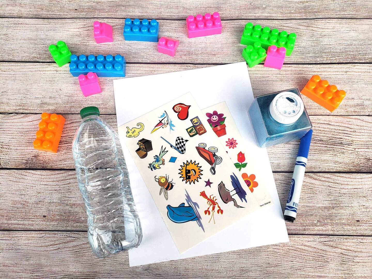 An empty plastic water bottle is laid out on a table alongside plastic blocks, stickers, a sheet of paper, a marker and a glitter container.