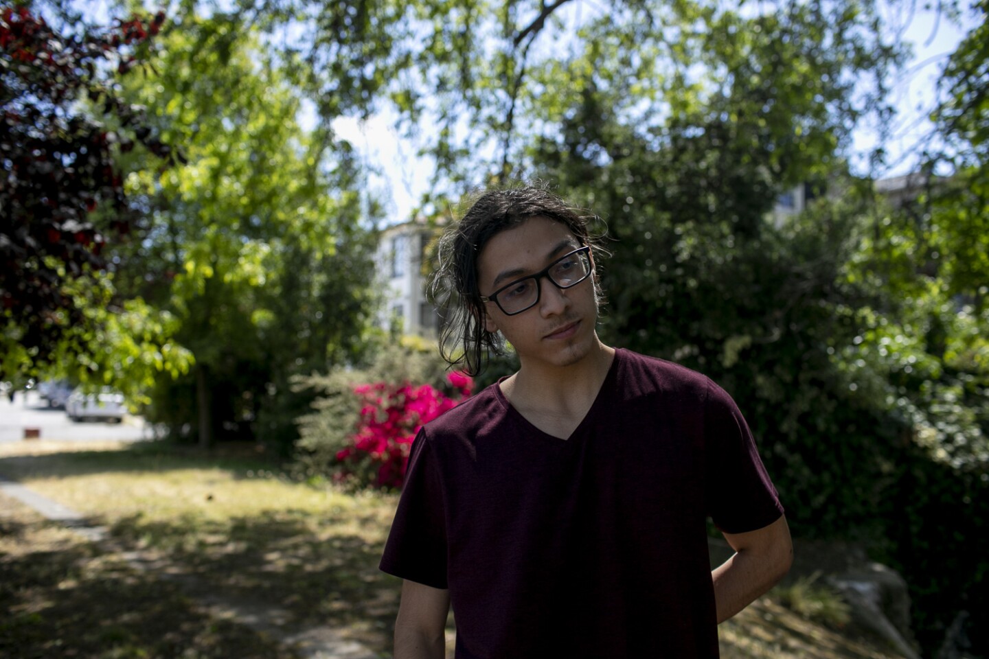 A young person in a dark red t-shirt, long, black hair, a small, trimmed beard and glasses stands outside near some greenery and looks to their left.