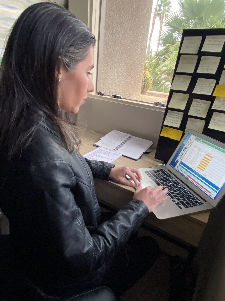 A woman in a black jacket and long gray hair types on a laptop.
