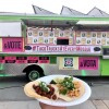 A lime green taco truck with bright pinks signs that read, "Vote" in Spanish, "Vota," in Arabic and in Chinese. In the foreground, someone holds up a styrofoam plate topped with two tacos.  