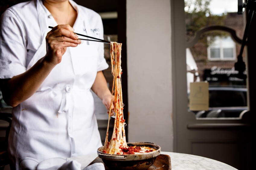 Chef Jenny Kwak is wearing a white apron and pulls the melted cheese out of a hot pot of dukboki fundido sitting on a marble table.  