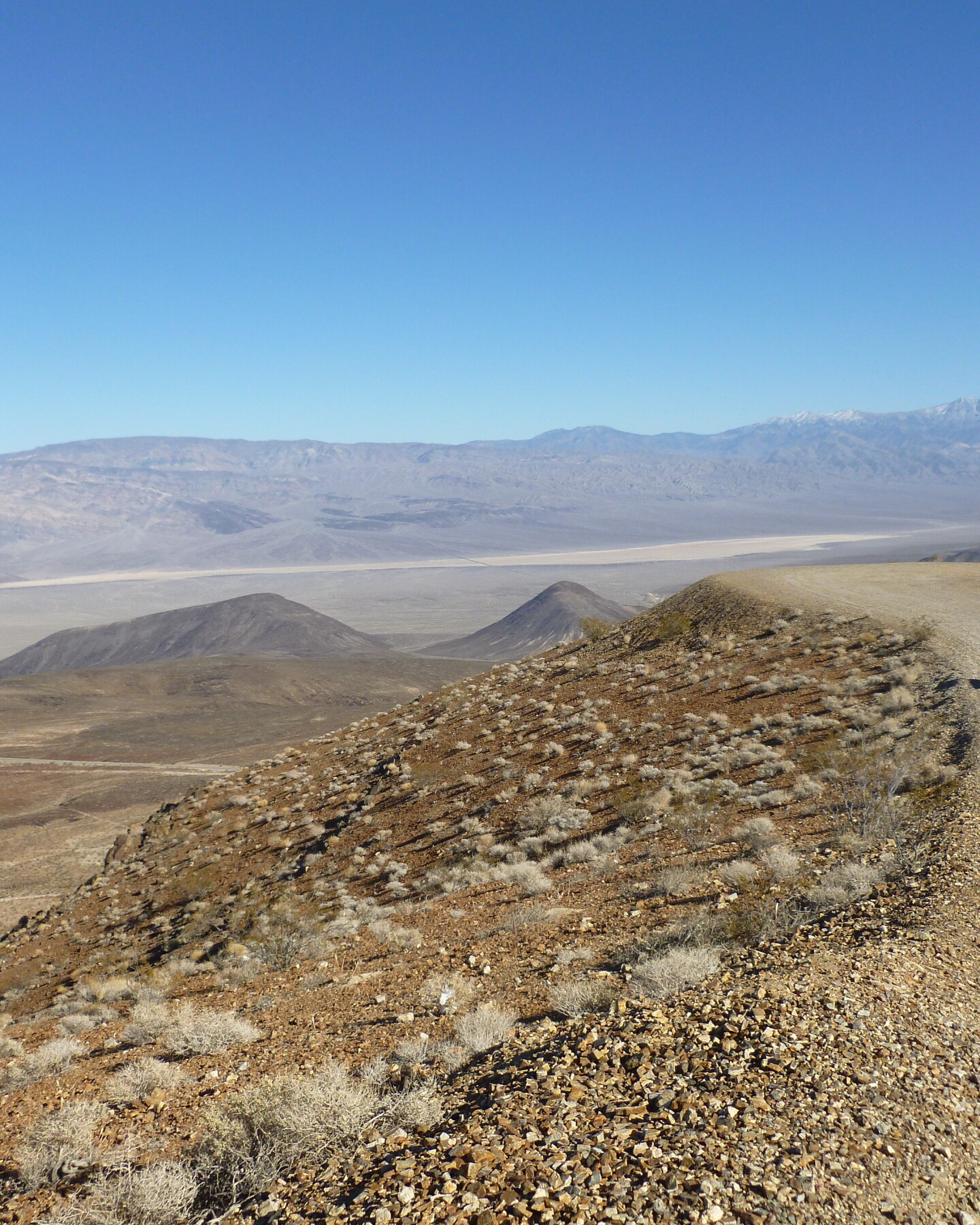 A silver jeep sits on a dirt path with a wide view of Death Valley behind it.
