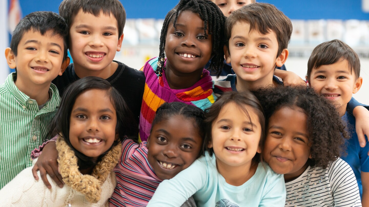 A group of diverse kids smile in this portrait. They are stacked on top of each other while cuddling in close and showing how happy they are.