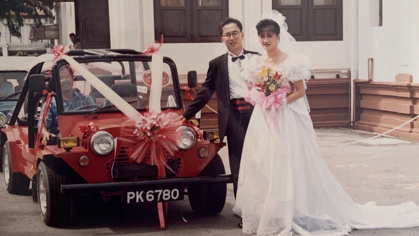 A bride and groom stand together and smile next to a little red car with a pink ribbon on it.