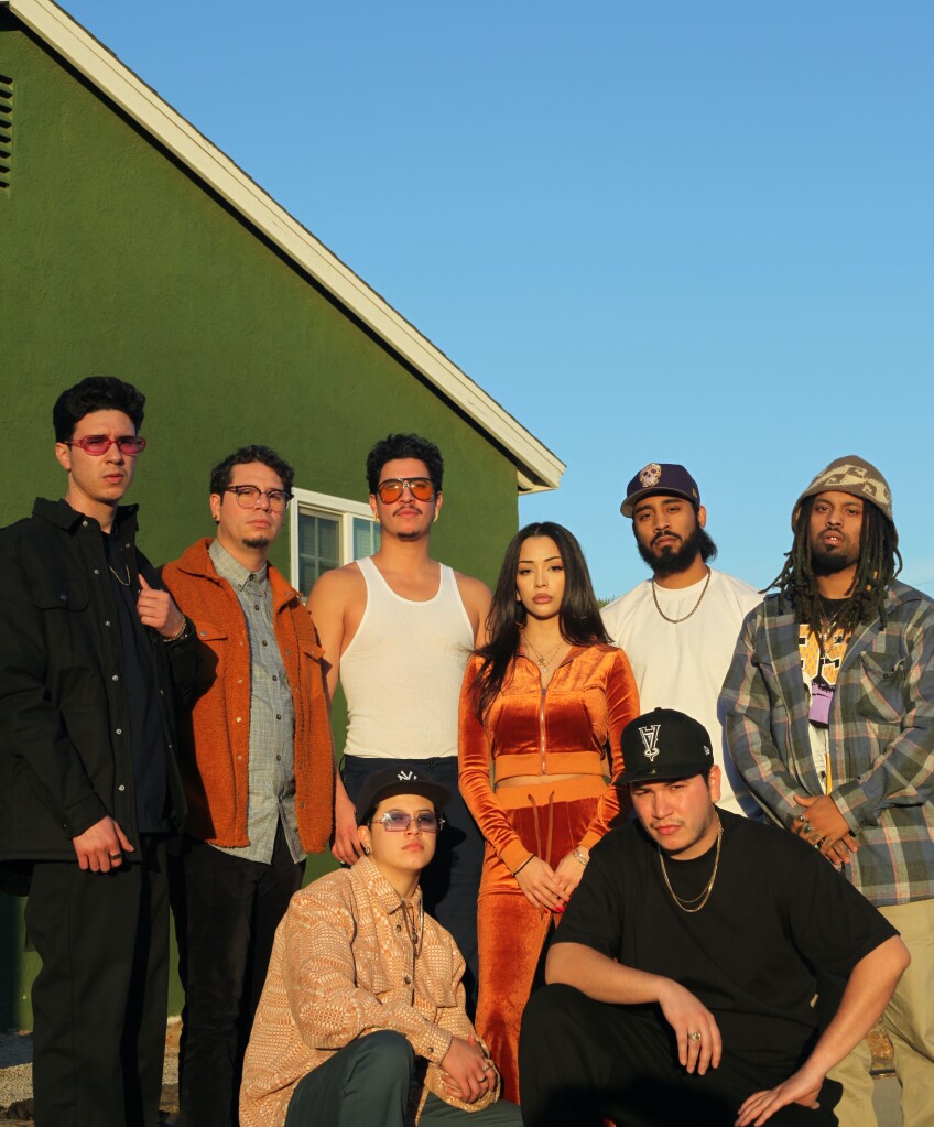 A group photo of Anaheim-based hip hop group Weapons of Mass Creation. They are all posed in front of a green house and against a clear blue sky. They are lit in a golden light from the sunset. 