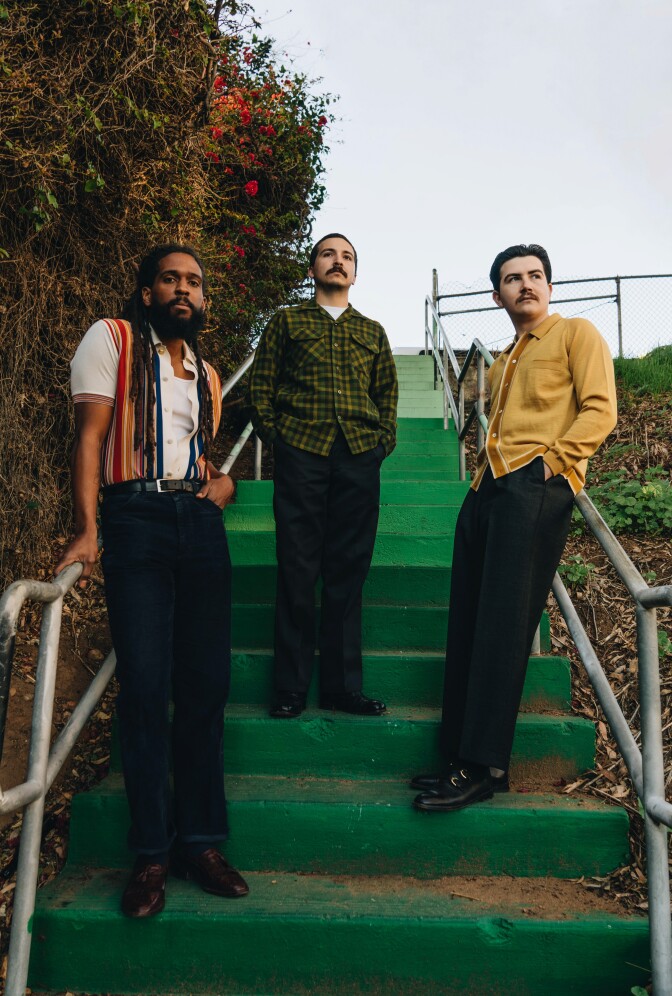 Three men stand in a triangle formation on a set of green stairs outdoors. The man on the left is wearing a vibrantly colored striped vest and has a thick beard. The man in the middle has a mustache and wears a green plaid button up with his hands in his pockets. The man to the right wears a yellow button up and also has a mustache. The men to the left and right lean against the railing of the stairs. To the left of the stairs is a wall covered in foliage and vining.