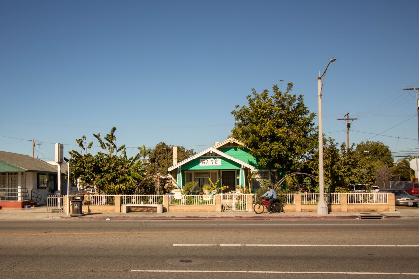 A teal house with a hand-painted sign that reads, "MAYE" stands behind a white wrought iron fence. The house is surrounded by tall, lush, green trees, produce, and shrubs. In front of the house's lot, on the sidewalk, is a biker on a red bike.