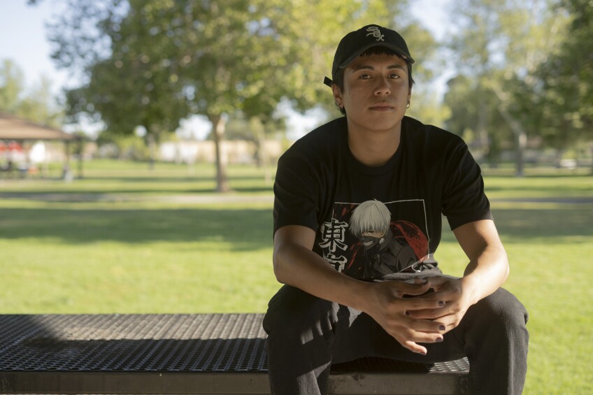 A young olive-skinned person wearing a black cap, grey painted nails and a black t-shirt with an Anime character on it looks at the camera with a very slight smile. He is sitting on top of a table at a park.