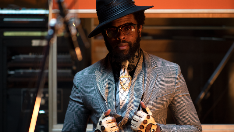 Adrian Younge poses facing the camera. He's wearing a plaid suit jacket and a silk black and white tie. He's also wearing a black brimmed hat tilted slightly to the left. Thick-framed glasses sit on his face and he's wearing white, fingerless gloves.
