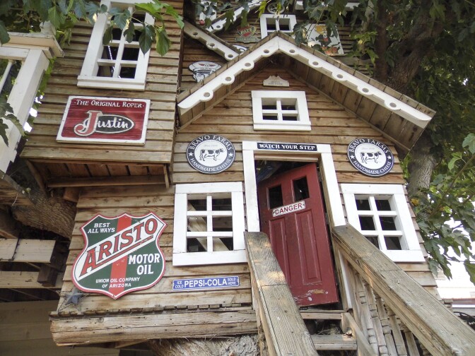 A treehouse made up of wood panels stands tall in the photo. A red door into the treehouse is slightly ajar with a "DANGER" sign attached to it. Various vintage signs decorate the exterior of the treehouse. An "Aristo" motor oil in the shape of a shield is to the left of the door. A "Pepsi-Cola" sign designed like a street sign is directly under a window. Two circular signs read, "Bravo Farms" on either side of the red door.
