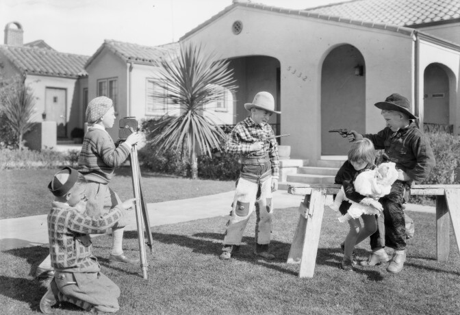 Children playing, some dressed up as cowboys and others posed like cameramen in front of a well-appointed home.