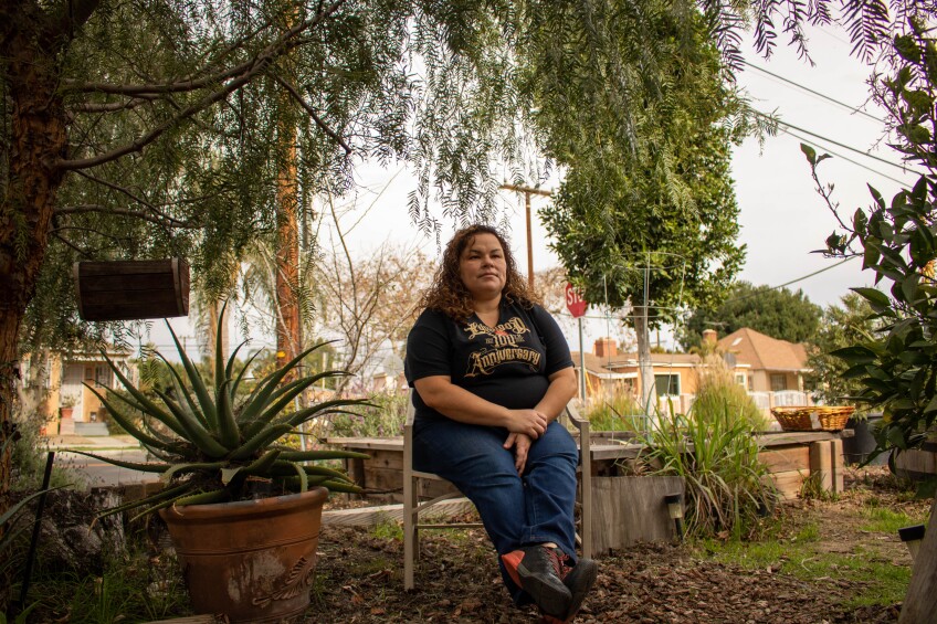 A woman wearing a black t-shirt and blue jeans sits on an outdoor chair underneath a big tree. She's surrounded by plants and gardening beds. Her hands are folded and she looks to the side.