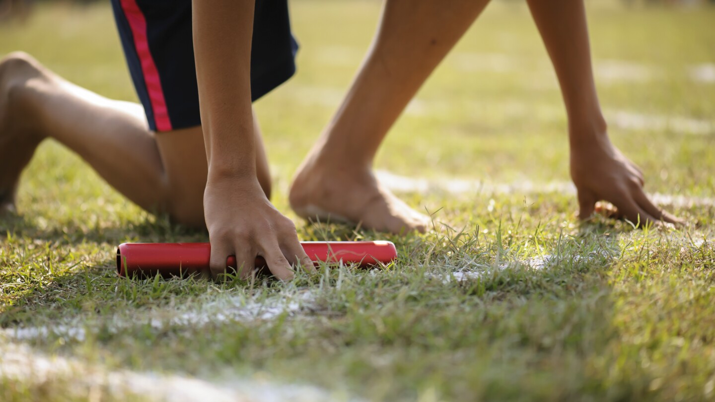 The athlete holding the relay race wood baton, is kneeling down on the track, preparing to start the first leg of a relay race. family math relay activity olympics