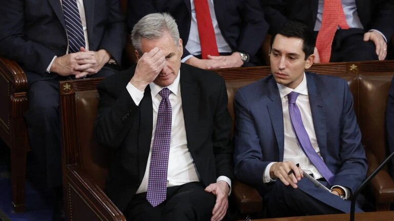 A white man wearing a dark suit and tie holds his right hand up to his forehead as he looks down while sitting in a leather-upholstered wooden chair