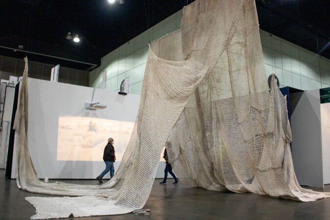A large net woven with a crochet-style technique is draped over the exhibition space. The net is woven with a plastic beige-colored material. Behind the net is a projection on a white gallery wall. The projections feature scenes of melting ice landscapes.