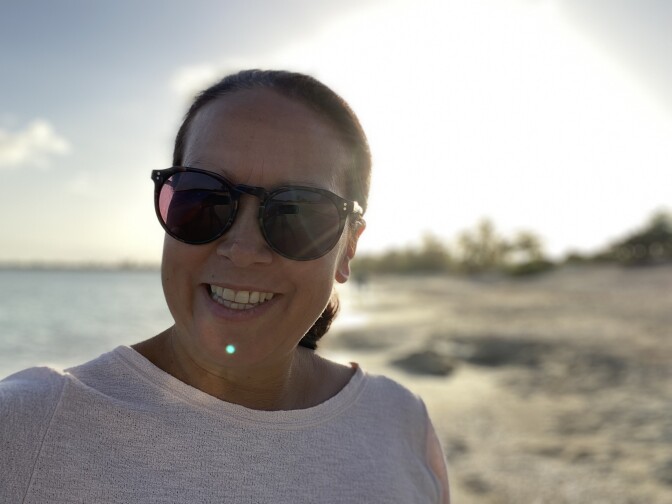 A woman in sunglasses smiles with a background of a beach