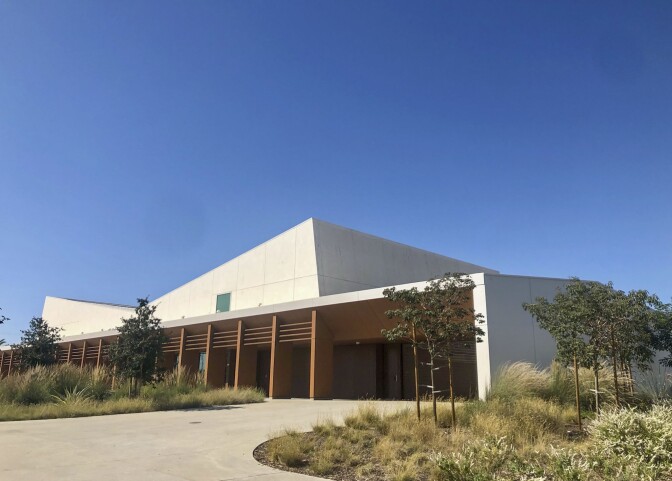 An angular building stands against a bright blue sky. Landscaped shrubs and trees stand in front of the concrete building.