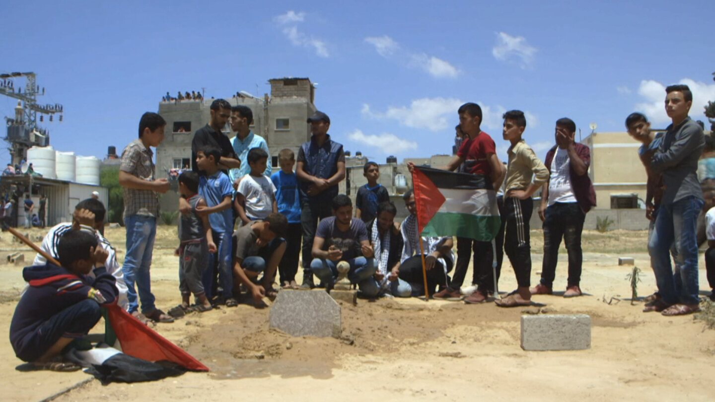 A group of men and boys crouch in a field around a Palestinian flag. | Still from "Witness" episode "Gaza: Between Fire & Sea"