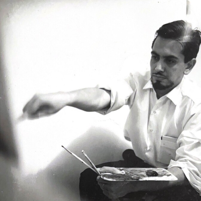 An old black and white photo depicts artist J. Sergio O’Cadiz Moctezuma seated with a palette holding paint in one hand and a paintbrush in the other. Only the back corner of the canvas he's painting on can be seen. O'Cadiz is young in the photo with dark hair slicked to the side and he's wearing a white button-up collared shirt. His face looks focused on his task.