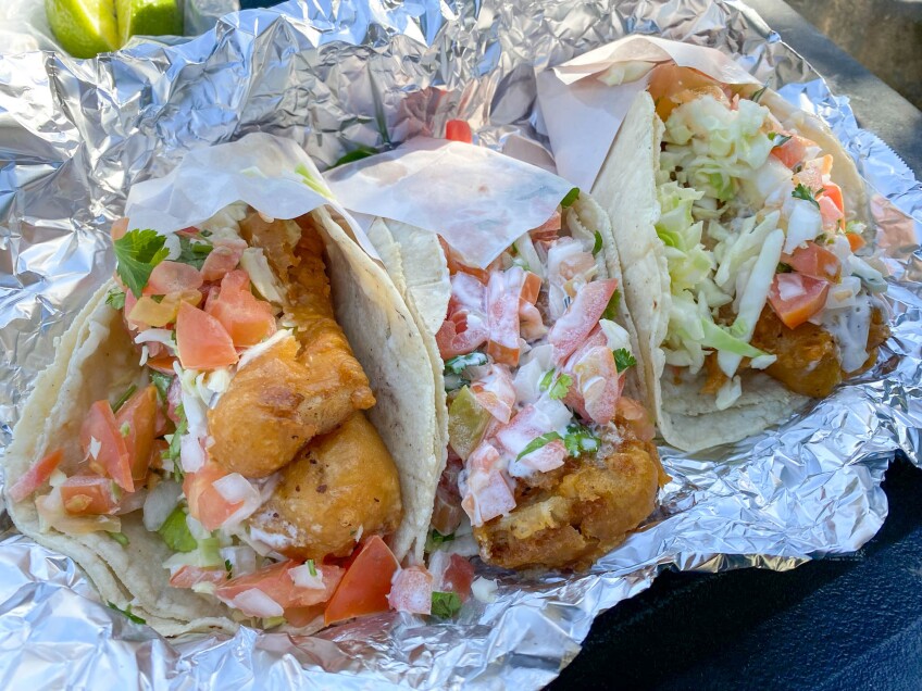 Three fish tacos sit together, nestled in tin foil. Each taco is filled with a golden crispy beer battered fried fish and topped with cabbage, a white crema sauce and diced tomatoes.