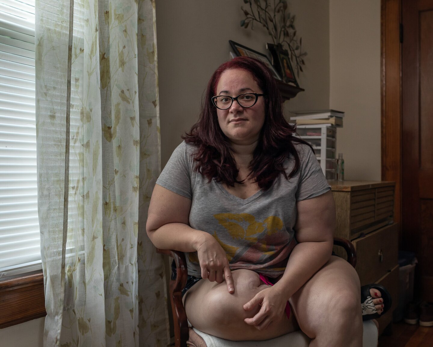 Monica sits at a chair next to a window lighting her face. She's sitting with one leg folded on the chair.