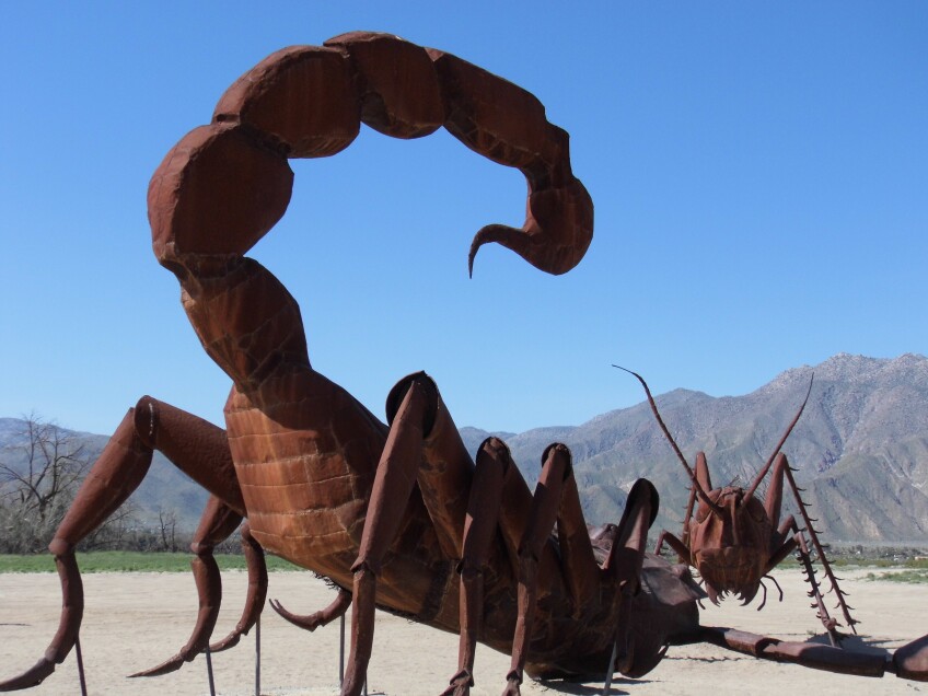 A red iron sculpture of a giant scorpion and a giant cricket/grasshopper stand in a desert landscape. 