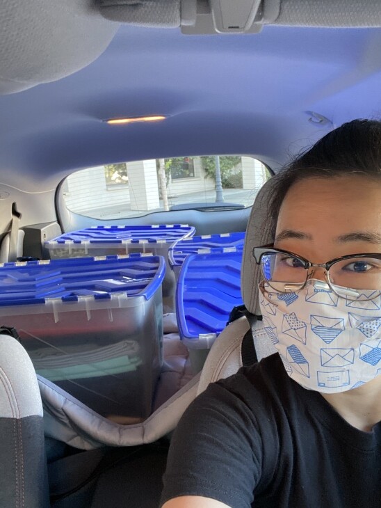 A face masked, bespectacled woman takes a selfie in her car, which contains materials to make cloth face masks.