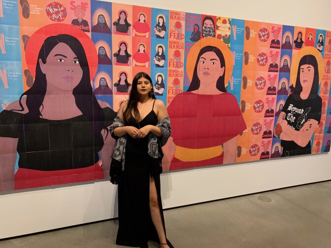 Adriana Arriaga wears a black maxi dress with a shawl over her arms. She stands before her colorful art piece consisting of several sheets of wheat paper arranged together to form a large art piece. The piece depicts three Xicana women, each with halos around their heads. They stand in front of small posters of various designs.