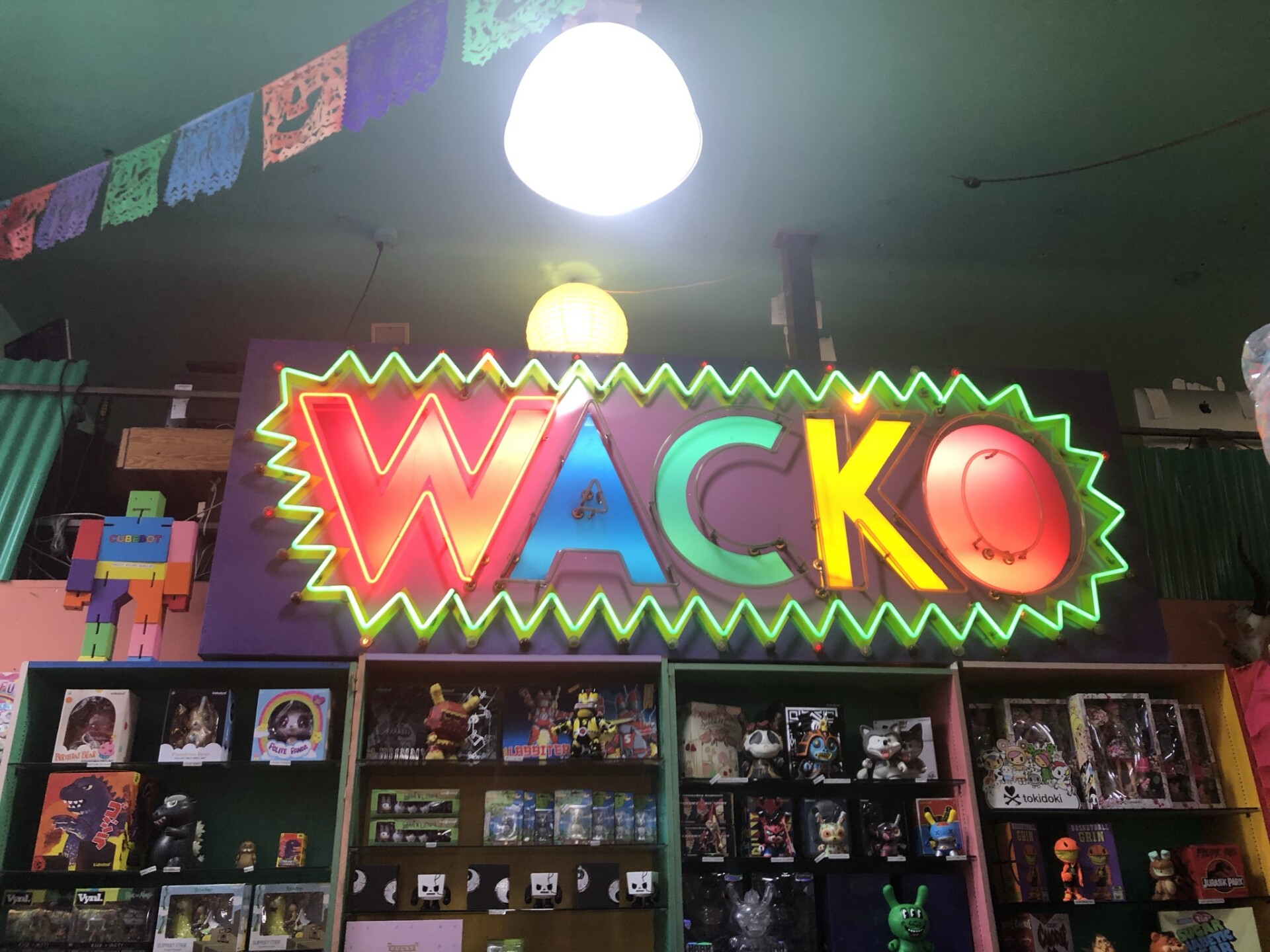 A neon sign that reads "WACKO" in different colors illuminates on the wall. The sign is large, taking up about half of the wall. Each letter is a different color, the W in red, A in blue, C in green, K in yellow and O in red. The sign in its entirety is outlined by a green, jagged neon trim. Below the sign are shelf displays of various ecclectic gifts like action figures and art. 