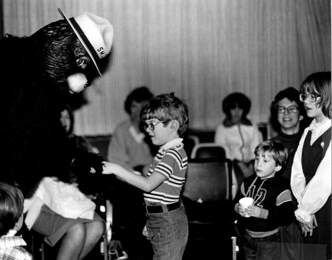 Smokey Bear shakes hands with a young boy in glasses and a striped shirt while other children watch.
