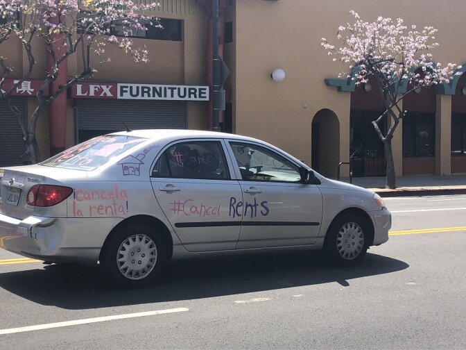 Car drives in rent relief protest, with paint on sides reading, "Cancela la renta" and "Cancel rents" | Karen Foshay