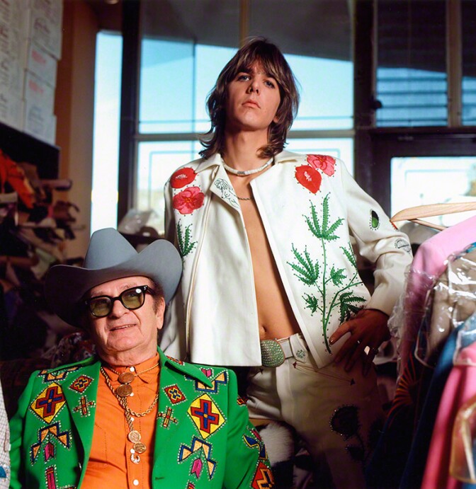 Gram Parsons (standing), adopting the rhinestone look of his country music heroes, in a personalized suit designed by Nashville's favorite tailor, Nudie Cohn (seated), at Nudie's Rodeo Tailors shop, Los Angeles, 1968 © Raeanne Rubenstein