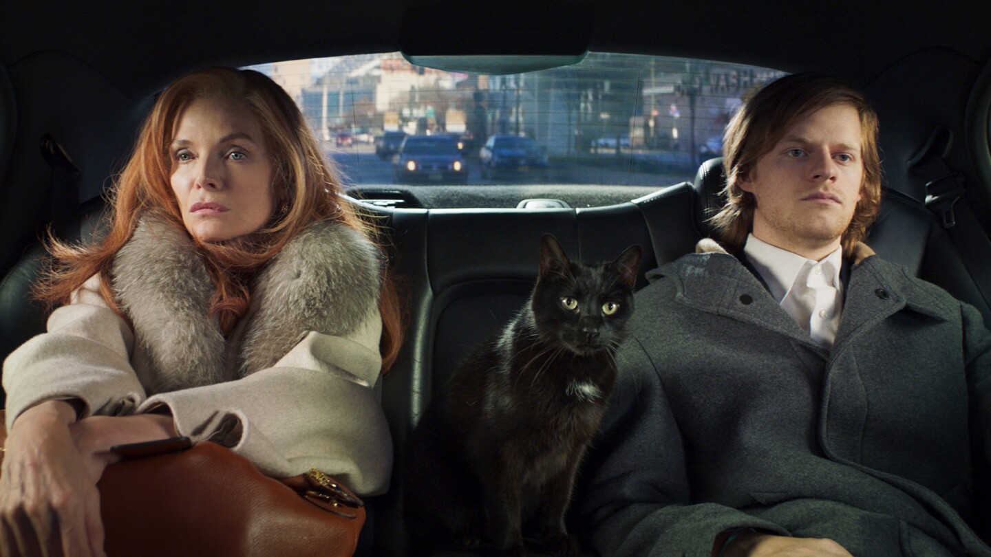 A woman and her adult son sits in the back of a car with a black cat between them.
