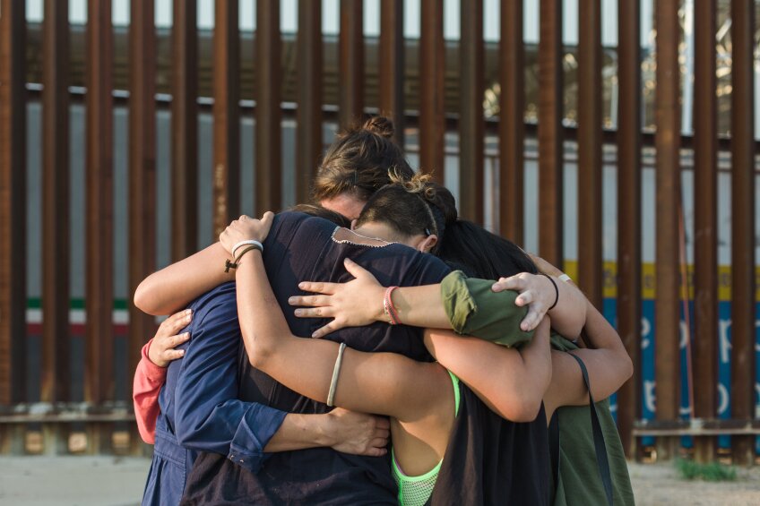 Four people in a tight hug by the U.S.-Mexico border.
