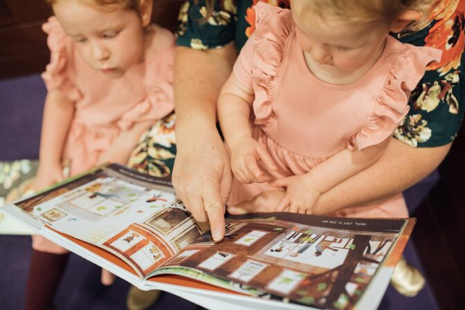 A woman reads over two little girls' shoulders who are wearing matching pink dresses as they hold a picture book.