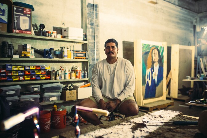 Rafa Esparza, who wears a white long-sleeved t-shirt and khaki shorts, sits on a stool in an art studio. Behind him is a large painting in progress and shelves filled with various art supplies.